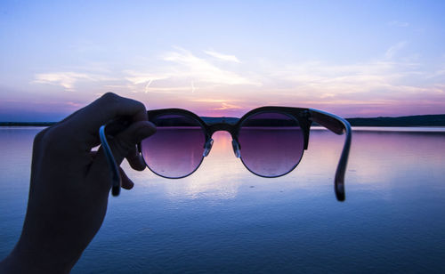 Cropped hand holding sunglass against lake during sunset