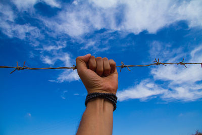 Cropped hand of man holding barbed wire against blue sky