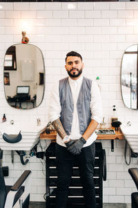 Self assured adult bearded male hairstylist in waistcoat looking at camera in barbershop
