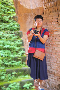 Portrait of woman with camera standing against brick wall