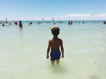 Rear view of boy standing in sea against sky on sunny day