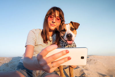 Low angle view of young woman taking selfie with dog through smart phone at beach against clear sky