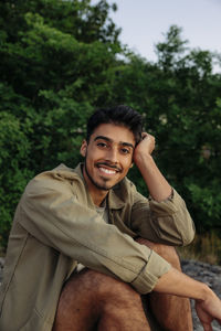 Portrait of smiling young man leaning head on elbow while sitting against plants