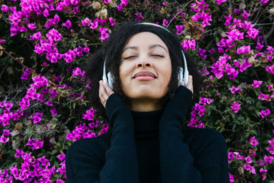 Content african american female in black wear listening to music in wireless headphones while standing near blooming bush with flowers