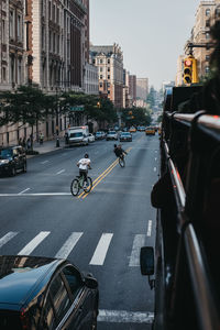 Rear view of men riding bicycle with cars on city street by buildings against sky