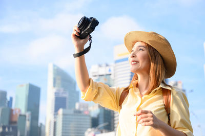 Portrait of smiling young woman holding camera against sky