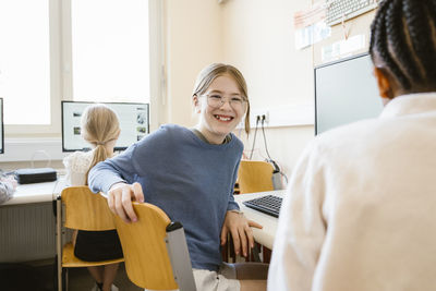 Portrait of happy female student sitting on chair with friends in computer class at school