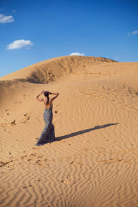 Brunette woman in a long leopard dress stands with her back in the desert at sunset. go everywhere