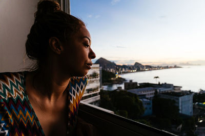 Side view of young woman looking out of a window from a bedroom to ipanema beach