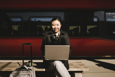 Portrait of smiling businesswoman with laptop sitting on bench in front of train at railroad station