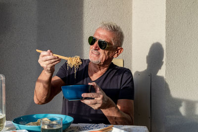 Attractive middle-aged man have fun while eating sitting on table laid chinese food in front of wall