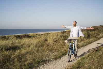 Carefree mature man with arms outstretched closing eyes while riding bicycle at beach