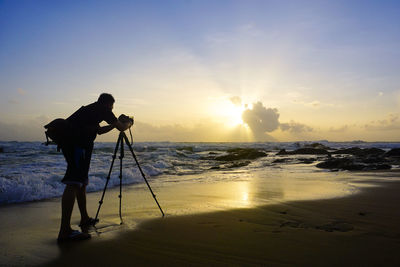 Man photographing at beach against sky during sunset