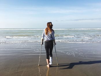 Full length of disabled woman with walking canes standing at beach against sky