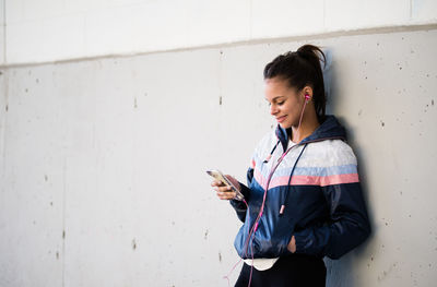 Young woman listening music through headphones while leaning on wall