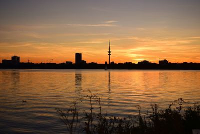 Silhouette factory by lake against orange sky