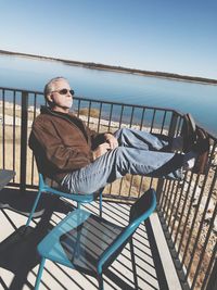 Full length of man relaxing against lake on chair during sunny day