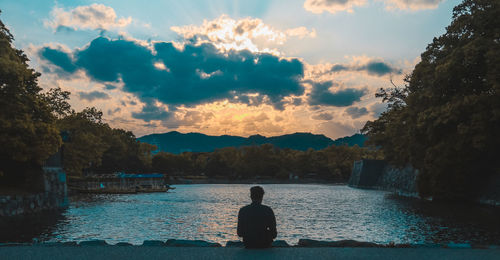 Rear view of man sitting by lake during sunset