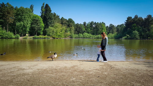 Side view of mother and son standing at lakeshore against trees