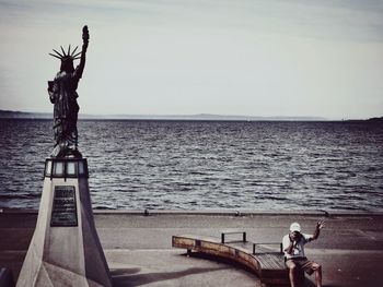 Man sitting on bench by statue of liberty replica at alki beach