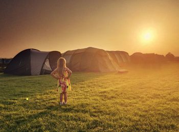 Rear view of girl with hands on hip standing on grassy field by tents against sky during sunset