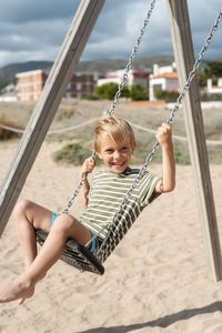 A happy boy on a swing on a sandy beach against the backdrop of apartment buildings and a stormy sky