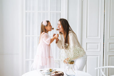 Funny little girl with mother young woman in white holding popcake in hands in bright living room 