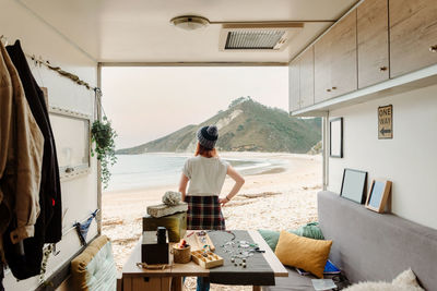 Back view of unrecognizable traveling female artisan standing near truck with handmade accessories on background of sea and mountains