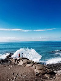 People fishing while waves splashing on rocks against blue sky