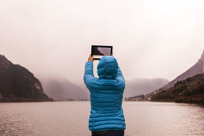 Rear view of woman photographing with digital tablet by lake during foggy weather