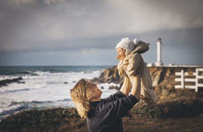 A woman is holding a baby near a lighthouse on the pacific coast
