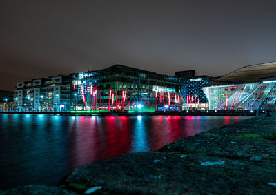 Illuminated buildings by river against sky at night