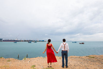 Rear view of couple holding hands while standing at beach against cloudy sky