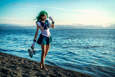 Full length of young woman photographing while standing in sea against sky during sunset