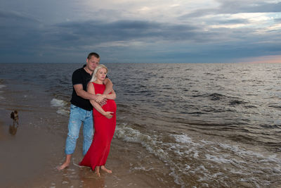 Pregnant couple standing at beach against sky during sunset