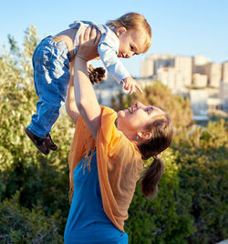 Side view of cheerful mother holding son against trees