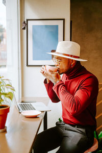 Side view of trendy african american male entrepreneur sitting in cafe and enjoying fresh coffee during break at remote work