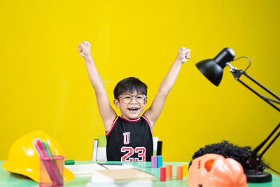 Full length of happy boy with arms raised against yellow wall