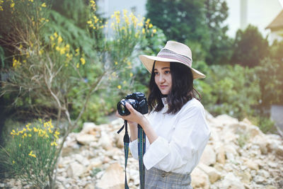 Young asian woman in white shirt and hat in a park, smiling, holding camera. outdoor portrait.