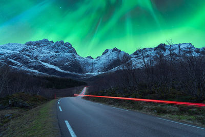 Breathtaking scenery of rocky mountain range covered with snow and empty asphalt road under night sky with purple and green aurora borealis in norway