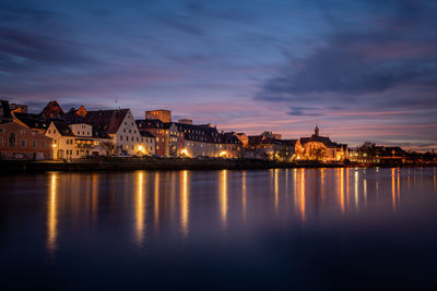 Illuminated buildings by lake against sky at sunset