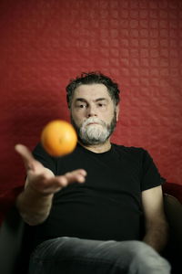 Man playing with orange while sitting against red wall