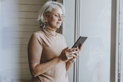 Mature woman with gray hair using tablet pc by window at home