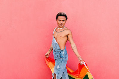 Portrait of gay man holding rainbow flag against pink wall