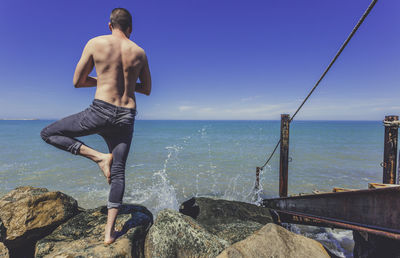 Rear view of shirtless man doing yoga on rock at seashore against clear blue sky