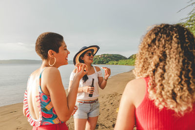 Group of cheerful young ethnic people chatting friendly and laughing while eating pizza and drinking beer together on sandy seashore in costa rica