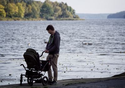 Man holding baby carriage while standing against lake