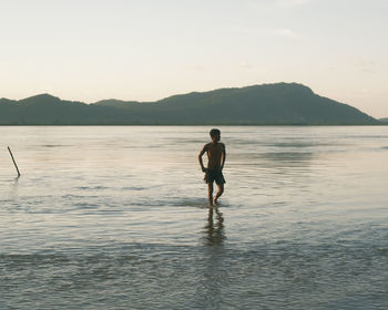 Rear view of man standing in sea against sky during sunset