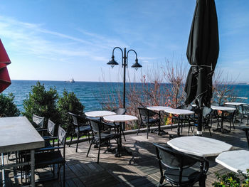 Empty chairs and tables at restaurant by sea against sky