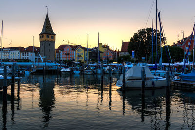 Sailboats moored in harbor against buildings in city during sunset
